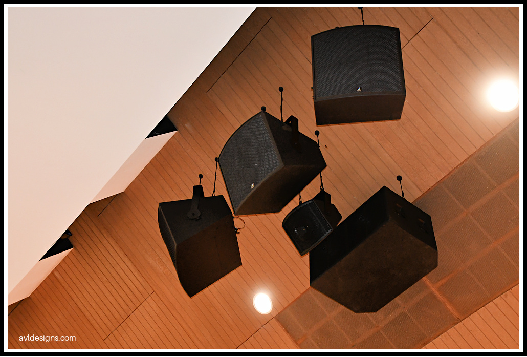 Four large black square speakers suspended from a wood-paneled ceiling in a performance space.