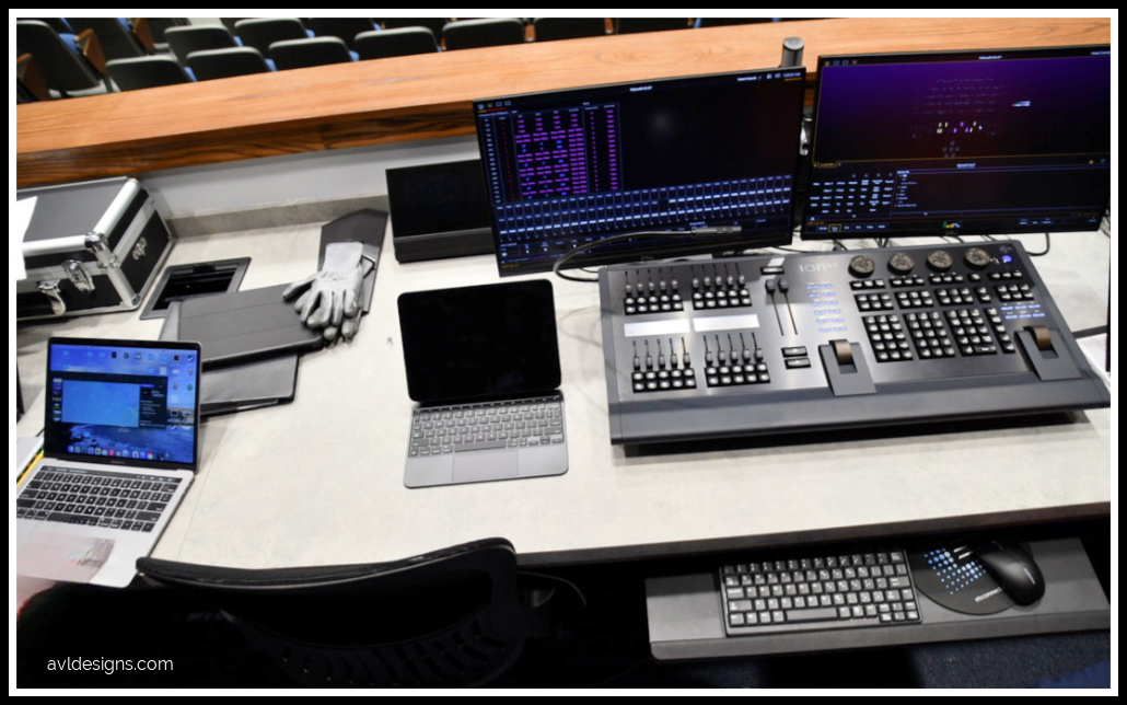 Control workstation with multiple monitors, keyboard consoles, and laptops on a conference-style desk, including a tablet and a pair of gloves nearby.