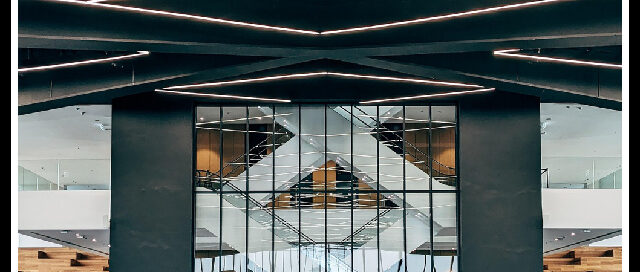 Interior of a modern lobby with dark square pillars, geometric LED ceiling lights, and a glass doorway reflecting on a glossy tiled floor.